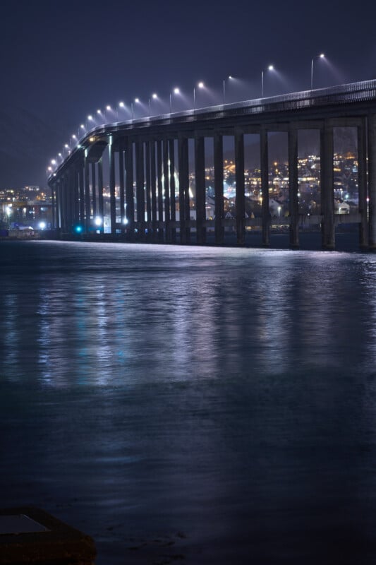 A long, illuminated bridge stretches over a calm body of water at night, with city lights glowing in the background. The reflections of the lights shimmer on the water's surface, creating a tranquil scene.