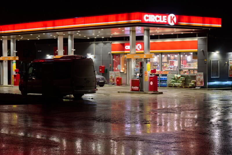 A Circle K gas station at night, illuminated by bright lights. A van is parked near the fuel pumps. The wet ground reflects the lights, creating a glossy surface. Inside, shelves stocked with various products are visible through the glass windows.