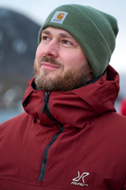 A bearded man in a red jacket and green beanie stands outdoors. He is looking upwards and smiling slightly. The background is blurry, suggesting a chilly, possibly mountainous environment.