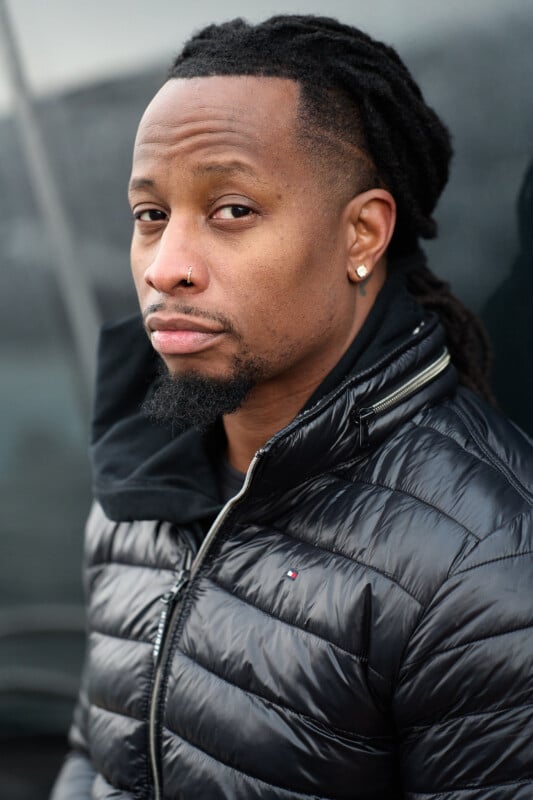 A person with long, braided hair and facial hair is wearing a black puffer jacket and a nose ring, looking directly at the camera. The background is a dark, out-of-focus surface.