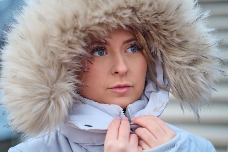 A person with blue eyes and light makeup looks to the side while wearing a fluffy hooded jacket. The fur hood is framed around their face, and they are holding the jacket's zipper with both hands. The background is slightly blurred.