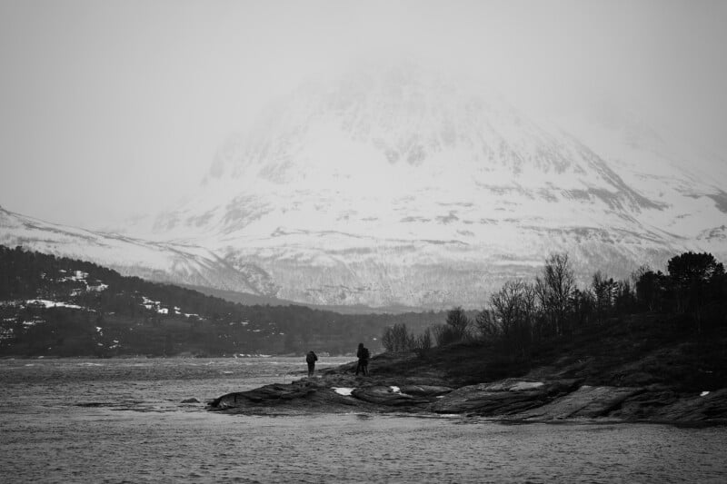 Black and white image of two figures standing on a rocky shoreline, facing a vast body of water. Snow-capped mountains loom in the background under a cloudy sky, creating a serene and atmospheric scene.