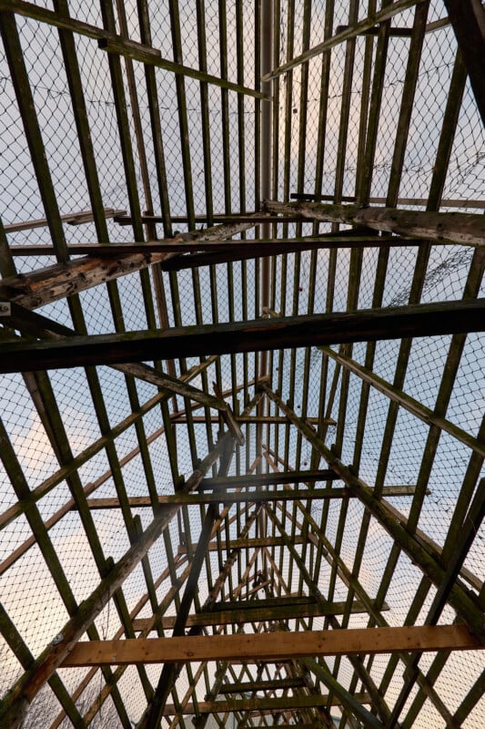 View from beneath a wooden and metal lattice structure resembling a roof, with a background of blue sky and clouds visible through the gaps. The geometric pattern adds depth and perspective.