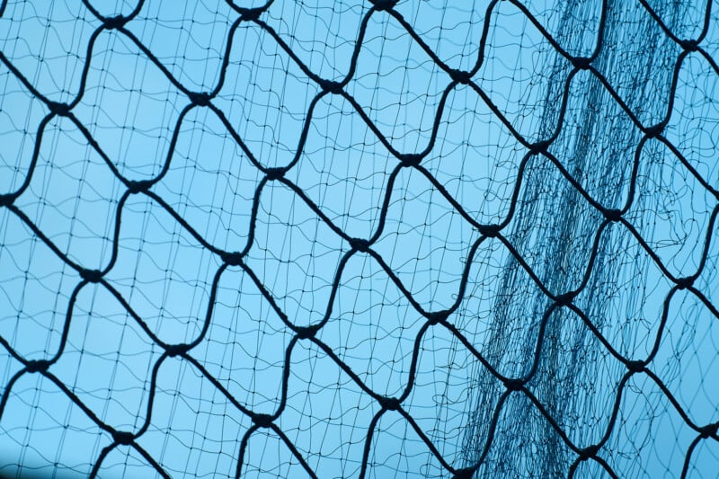 Close-up of a twisted wire fence against a clear blue sky. The crisscross pattern forms diamond-shaped spaces in the design, with the sunlight subtly highlighting the wire texture.