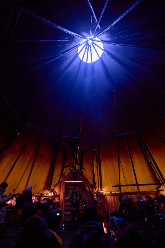 A dimly lit gathering inside a large tipi with a round opening at the top, casting blue light. People are seated around the edges, while others stand in the center, surrounded by a warm glow from small fires.