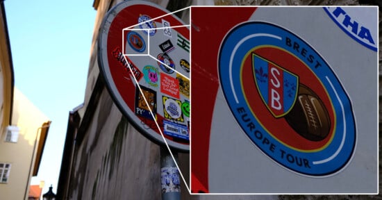 Close-up of a street sign covered with various colorful stickers. The sign is red with a white circle, partially visible, mounted on a metal pole. Stickers include a logo with overlapping letters "SB" and text "Brest Europe Tour.