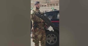 A soldier in camouflage uniform and beret stands guard, holding a large black and white weapon, next to a black vehicle with a partially visible tire. An outdoor urban setting is in the background.
