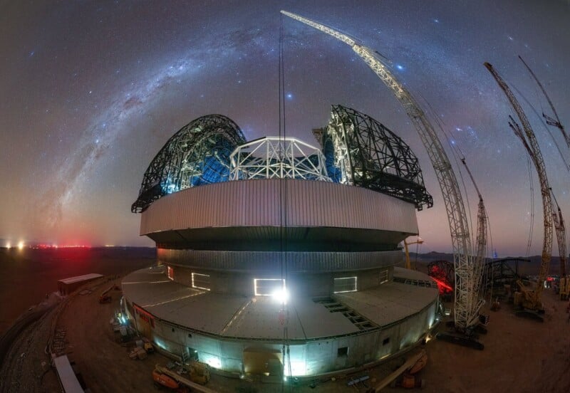 A large telescope under construction at night, surrounded by cranes and machinery, with the Milky Way and starry sky arching above in the background.