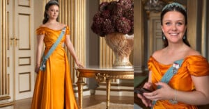 A young woman in an orange satin gown with a blue sash and tiara stands in an elegant room with gold accents, a floral arrangement, and holds a phone, smiling.