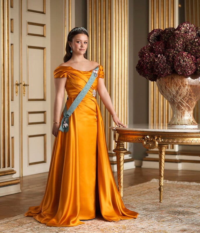 A young woman wearing a golden off-the-shoulder gown, blue sash, and tiara stands beside an ornate table with a large bouquet of dark flowers in an elegant, gold-accented room.