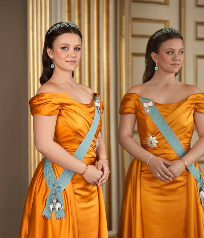 A young woman in an elegant, off-shoulder orange gown and tiara stands beside a mirror. She wears a blue sash with medals and jewelry, and both her and her reflection pose gracefully in a lavish room with gold accents.