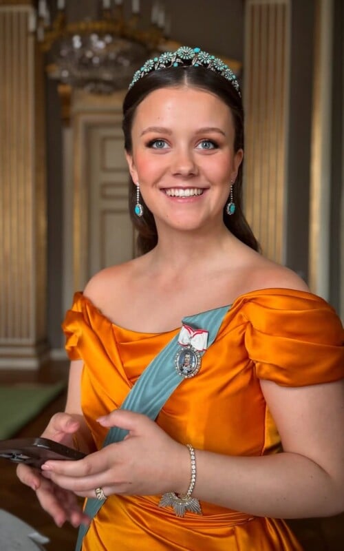 A smiling woman in a formal orange gown and teal sash stands indoors, wearing a jeweled tiara and earrings, with her hair styled neatly. She holds a phone and stands in an elegant room with columns and a chandelier.