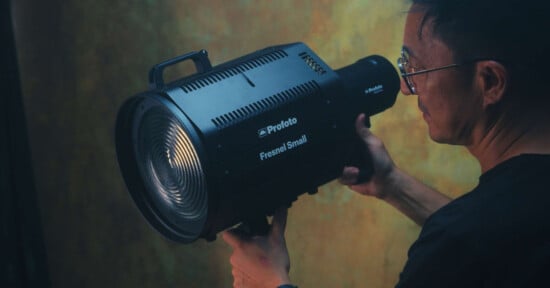 A person wearing glasses holds a Profoto Fresnel Small lighting unit, preparing to adjust or position it. The background is dimly lit with a mottled pattern, creating a dramatic and focused atmosphere.