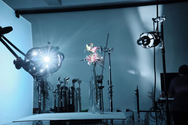 A photography studio setup shows bright lights and equipment focused on a vase of flowers on a table; a person sits at a computer on the right, partially out of frame.