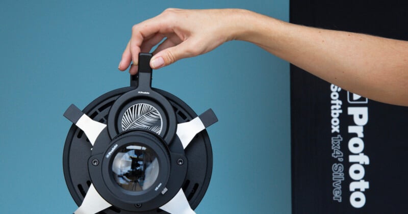 A hand adjusts a Profoto photography light modifier with a patterned insert, in front of a softbox labeled "Profoto Softbox 1x4' Silver" against a blue background.