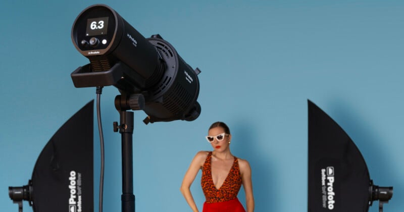 A studio lighting setup with two softbox lights and a strobe flash in the foreground, with a woman in sunglasses and a red patterned dress posing in front of a blue background.