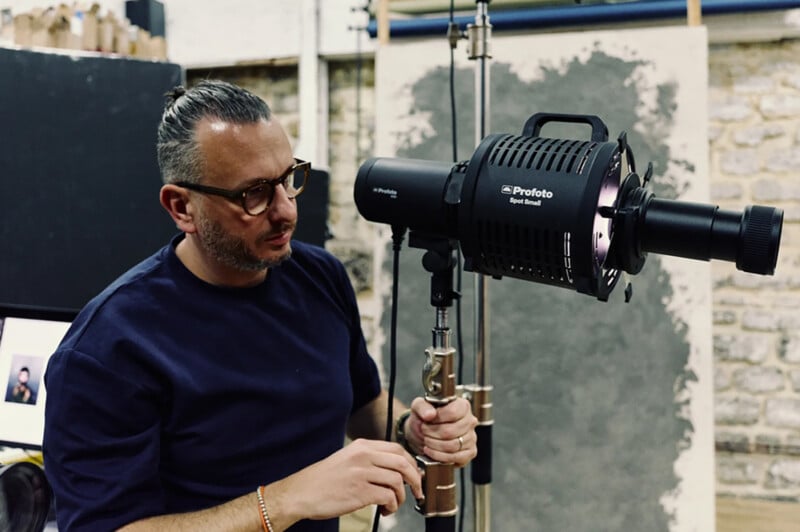 A man wearing glasses adjusts a large studio light on a stand in a photography studio with exposed brick walls.
