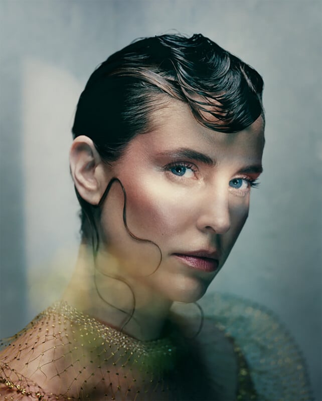 A close-up portrait of a woman with slicked-back dark hair and loose waves framing her face, wearing netted golden fabric, with soft lighting and a blurred, dreamy background.