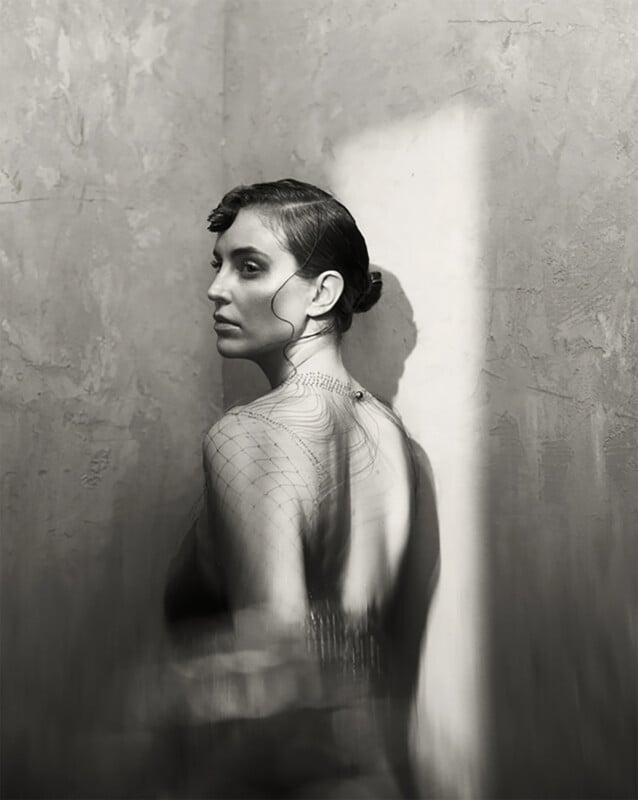 A woman with her hair in a low bun poses in profile, wearing a dress with an intricate back detail. Dramatic lighting casts her shadow on a textured wall, creating a moody, artistic black-and-white portrait.