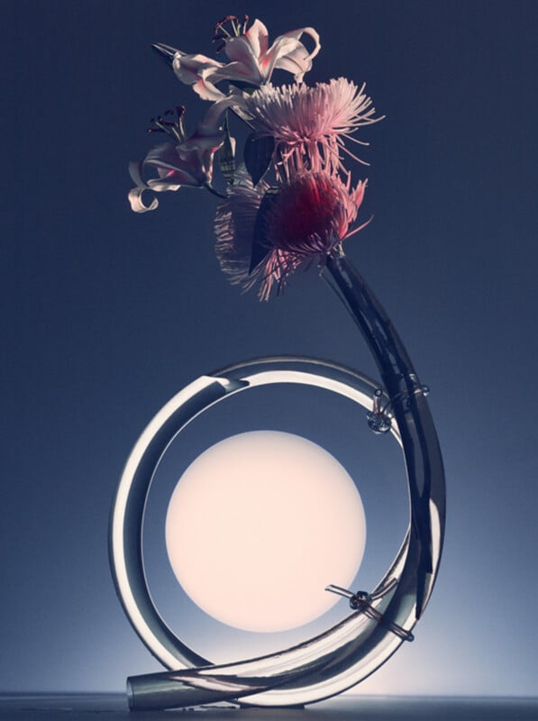 A sleek, curved glass vase holds pink lilies and chrysanthemums, set against a soft gradient background with a glowing white orb in the center, creating a modern, artistic composition.