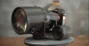 A paper model of a large camera lens mounted on a rotating display stand, with a smaller lens placed beside it. The background is a blurred gray wall, giving focus to the model.