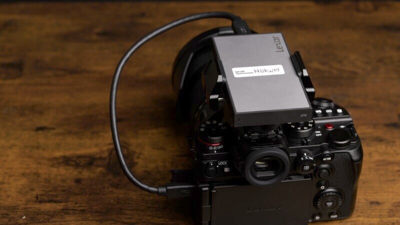 A professional camera is on a wooden surface with a Lexar storage device attached via a cable. The device has a label saying "Norway." The setup appears ready for storage or backup purposes.