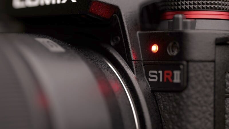 Close-up of a Lumix S1R II camera showcasing the lens mount area. A red LED light is illuminated near the lens release button, and the camera surface features a textured black finish with prominent model branding.