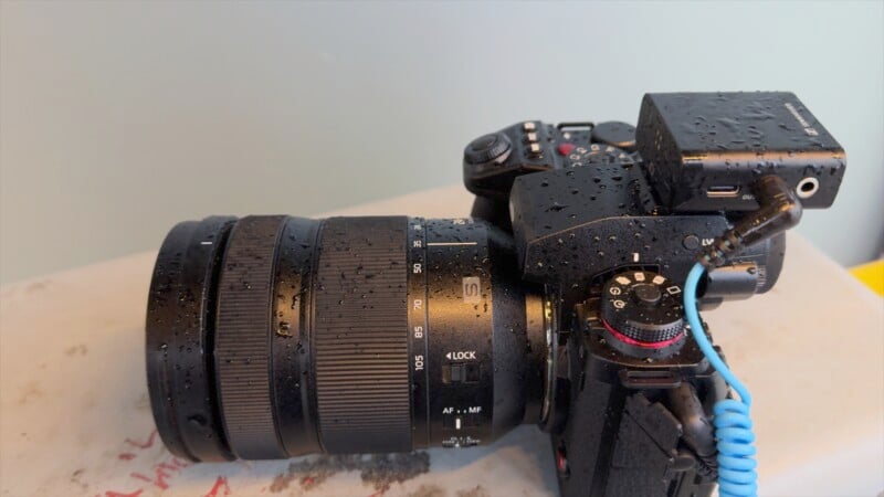 A black DSLR camera with a large zoom lens is wet with water droplets. It has a blue cable attached to the side, and it's resting on a beige surface with some red markings. The background is a light blue wall.