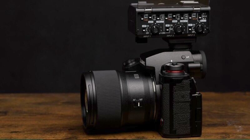 A black DSLR camera fitted with a large lens and an audio input attachment on top sits on a wooden surface against a dark background. The camera features multiple dials and buttons, highlighting its professional design.