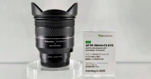A black Schneider-Kreuznach x LK Samyang camera lens is displayed on a clear stand next to a sign announcing the AF 14-24mm F2.8 FE lens, developed with Schneider-Kreuznach, and coming in 2025.