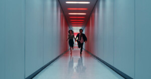 A woman and a man run side by side down a long, narrow hallway with pale walls and red lights on the ceiling, creating a dramatic and urgent atmosphere.