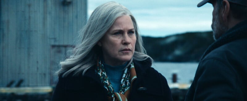 A woman with long gray hair and a serious expression stands outdoors by the water, wearing a dark coat and scarf, facing a man whose face is partially visible. The background shows a wooden building and a cloudy sky.
