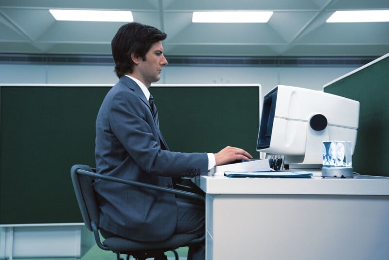 A man in a gray suit sits upright at a desk, typing on a vintage computer in a modern office with green dividers and fluorescent lighting. A cup with pens and papers is on the desk.