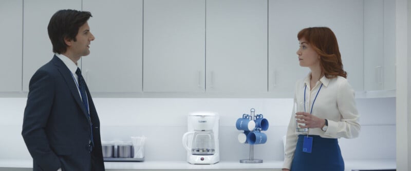 A man in a suit and a woman in business attire stand facing each other in a white office kitchen, with a coffee maker and blue mugs on the counter between them. The woman holds a glass of water.