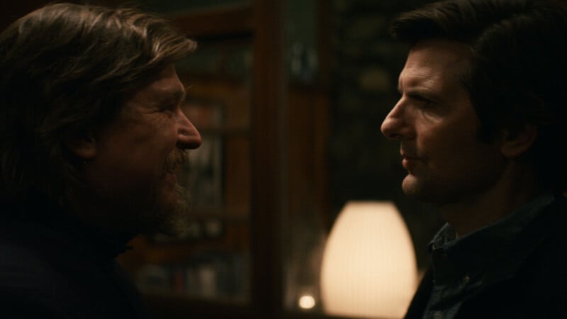 Two men stand face-to-face in a dimly lit room, one smiling with a beard and longer hair, the other serious with short dark hair. A glowing lamp and wooden furniture are visible in the background.