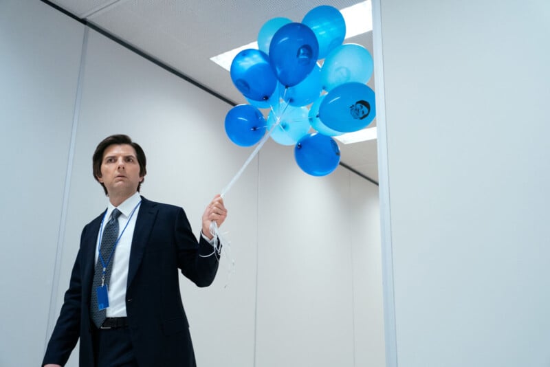 A man in a suit stands in a plain, white hallway holding a bunch of blue balloons. He looks to his left with a serious expression. The setting appears to be an office or institutional building.