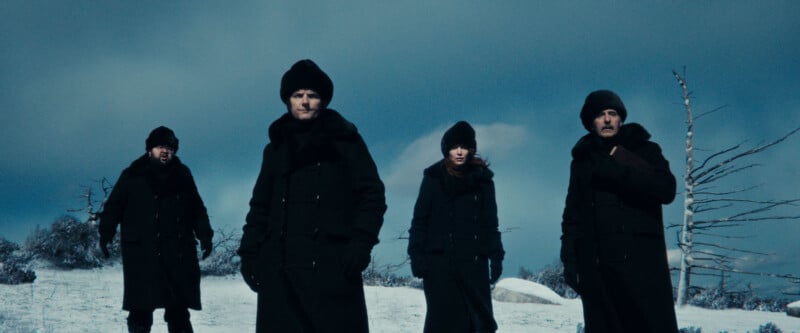 Four people in heavy black winter coats and hats stand in a snowy, barren landscape under a cloudy sky, with leafless trees and patches of snow in the background.
