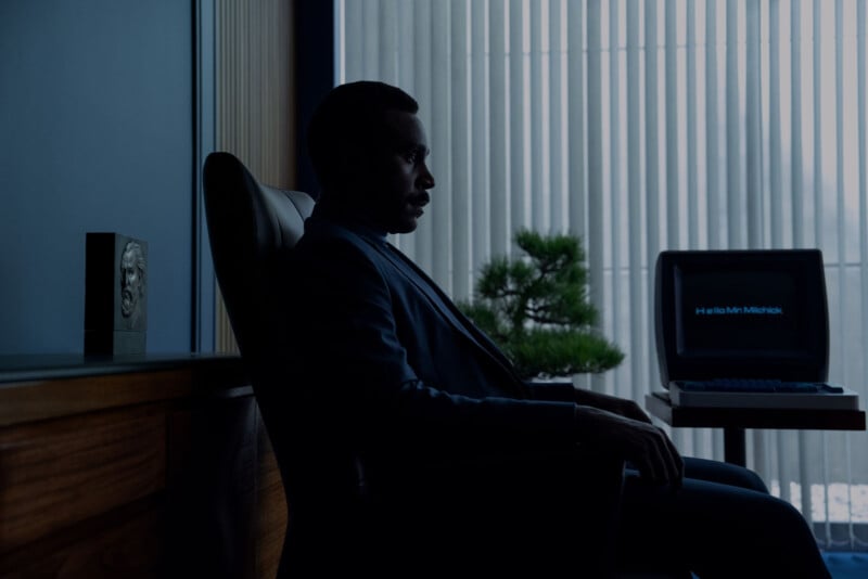 A man in a dark suit sits in a dimly lit office beside a desk with a vintage computer displaying “Hello Mr. Mishick.” Vertical blinds cover the window behind him, casting shadows across the room.