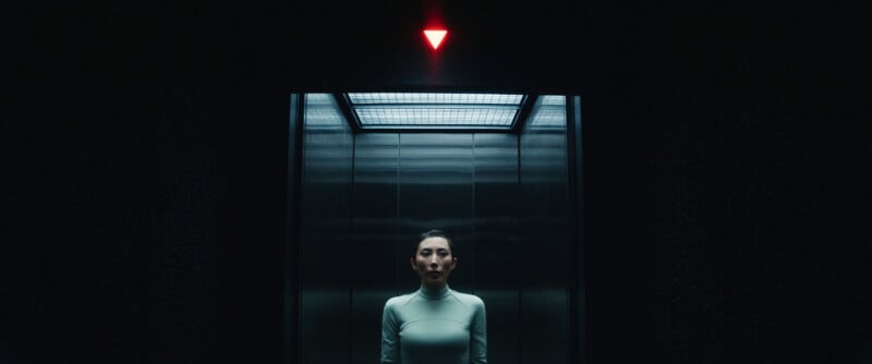 A woman stands alone in a dimly lit elevator with metallic walls. Above the elevator door is a glowing red downward-pointing triangle, casting a moody light over the scene.