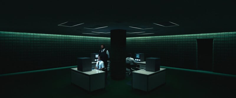 A dimly lit, futuristic office with green walls and four cubicles arranged around a central pillar. Three people sit at desks with computer monitors, while one stands, all illuminated by the monitors' glow.