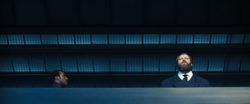 Two men stand behind a blue railing in a modern, industrial-looking building. One man, on the right, has a beard and wears a suit; the other man, on the left, is clean-shaven and looks sideways.
