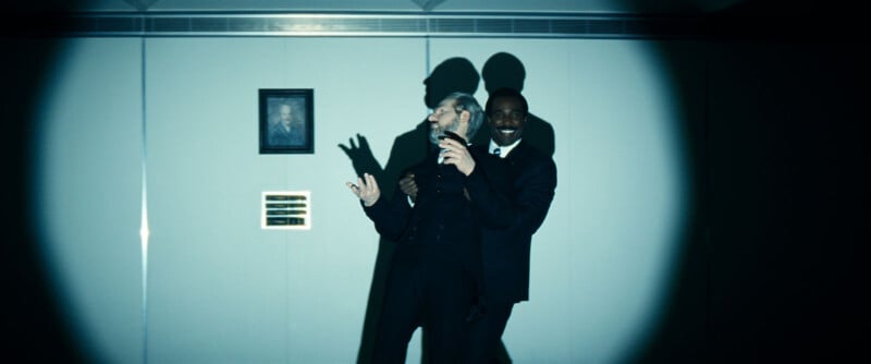 Two men in suits stand together in a spotlight against a plain wall; one holds a microphone and gestures, while the other stands behind, smiling. Their shadows are visible on the wall, along with a small portrait and plaque.