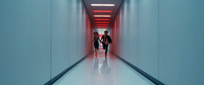 A man and a woman in business attire run down a long, narrow hallway with white walls and a white floor; red lights line the ceiling, creating a sense of urgency and tension.