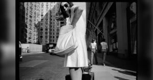 A woman in a light-colored dress stands on a city sidewalk, reaching into a large bag. She wears sunglasses and is surrounded by tall buildings, with two people walking away in the background.