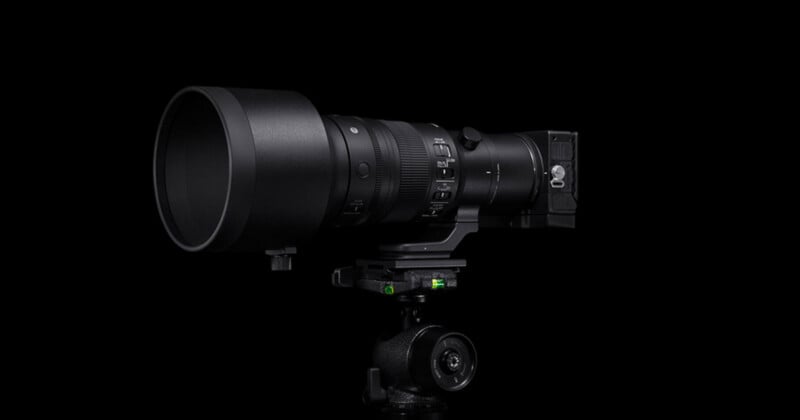 A large black camera lens with a wide hood is mounted on a tripod against a dark background. The lens features dials and switches, indicating adjustable settings, and appears suitable for professional photography.