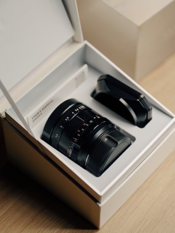 A camera lens and lens hood are neatly placed inside a white presentation box, alongside a user manual, on a wooden surface.