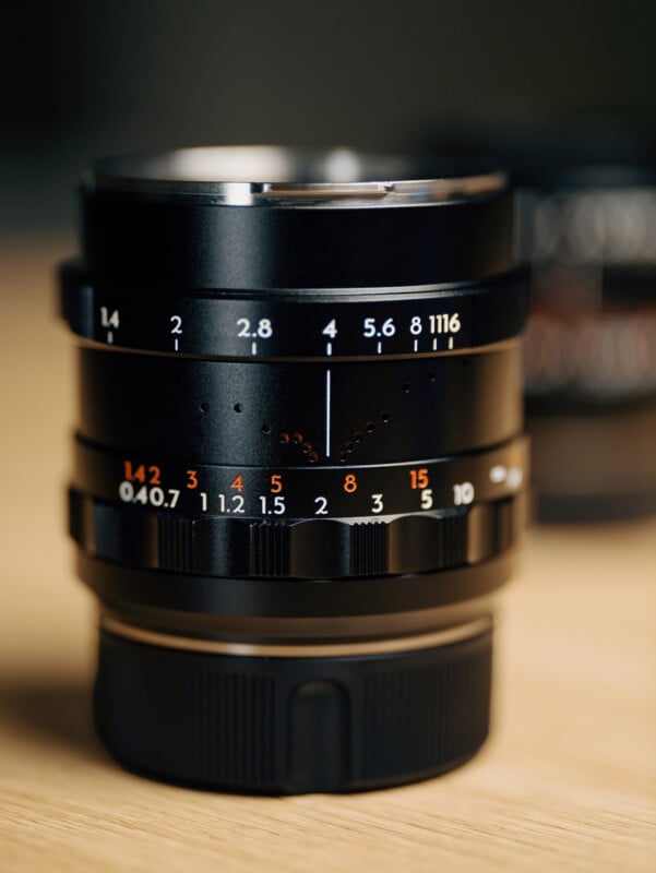 A close-up photo of a black camera lens with visible aperture and focus markings in white, orange, and red, resting on a light wooden surface with a blurred background.