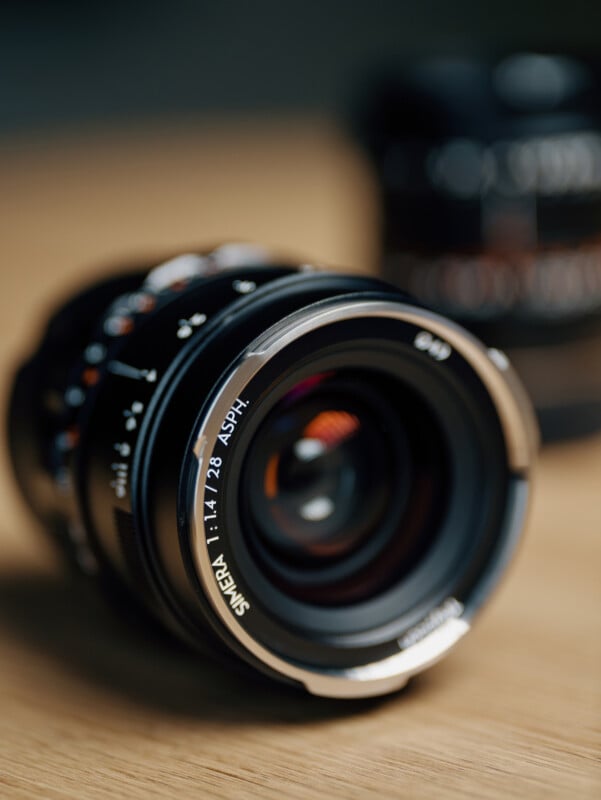 A close-up photo of a camera lens labeled "SMC 1:1.4/28 ASPH" resting on a wooden surface, with another blurred lens in the background.