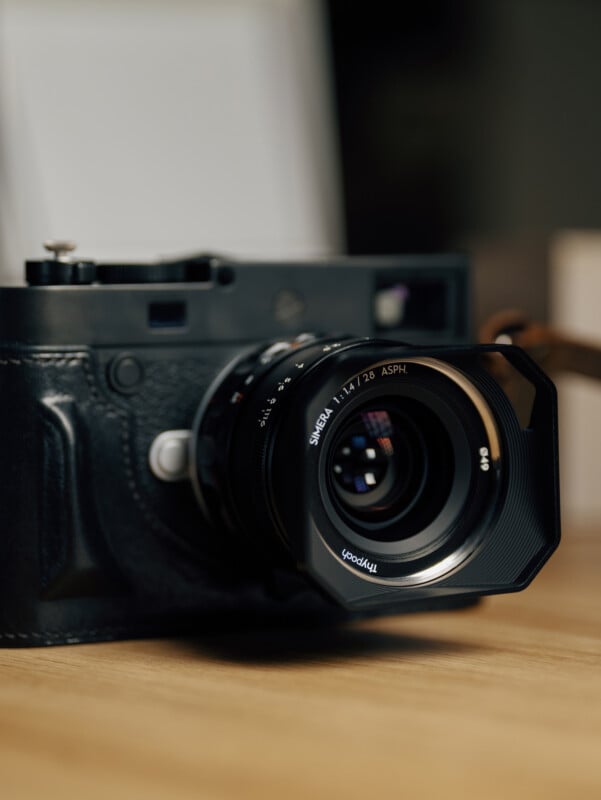 A close-up of a black camera with a large lens resting on a wooden surface, showing detailed texture and reflections on the lens glass.