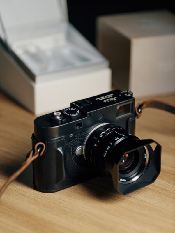 A black Leica camera with a leather strap sits on a wooden surface. The camera is in focus, with a blurred beige box and an open white box in the background.